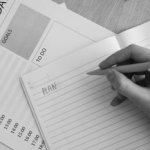 Black and white photo of hand writing 'plan' in notebook with planning materials around.